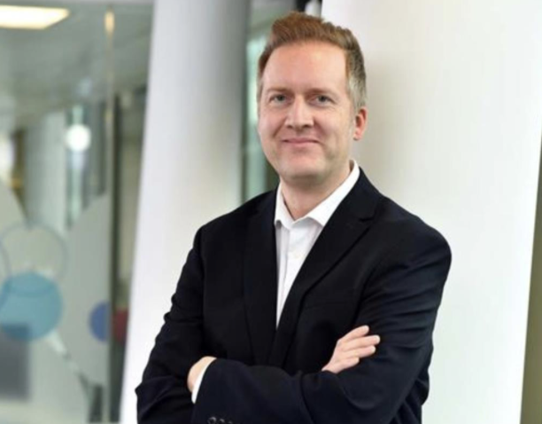 A man in a black suit with a white shirt standing with arms crossed in an office building. 