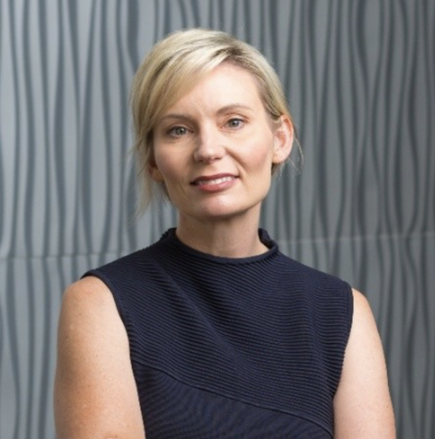 Portrait of a woman with blonde hair wearing a sleeveless black top, standing in front of a textured gray background.