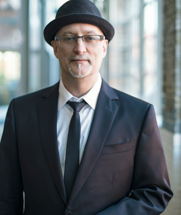 Man wearing a dark suit, white dress shirt, black tie, and black hat standing in a modern indoor space with large windows and natural light.