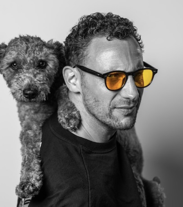 A man with curly hair and sunglasses with orange-tinted lenses has a dog on his shoulder, both looking at the camera against a plain background.