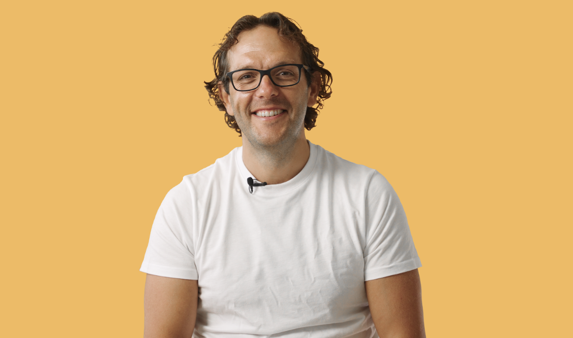 A smiling man with glasses and curly hair is wearing a white t-shirt and a lapel microphone, sitting against a yellow background.