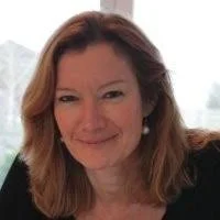 A woman with shoulder-length reddish-brown hair smiling, indoors with a window in the background.
