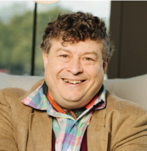 A smiling man with curly hair sitting on a couch indoors, wearing a plaid shirt, a colorful patterned tie, and a brown blazer.