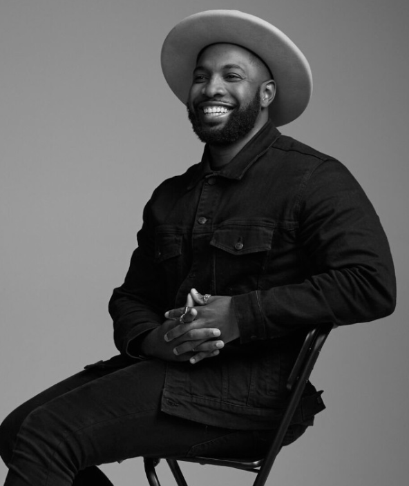 A smiling man wearing a wide-brimmed hat, a dark denim jacket, and dark pants, sitting on a chair with hands clasped, against a plain gray background.