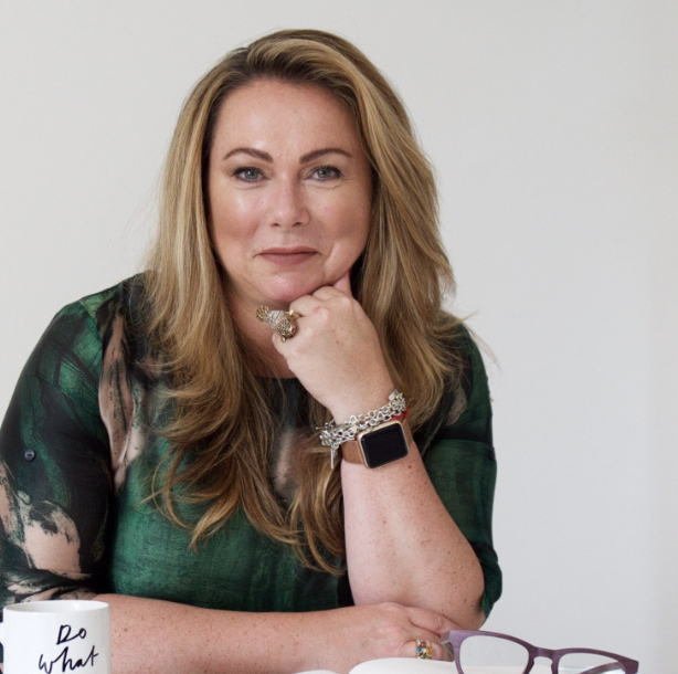 Portrait of a woman with long blonde hair wearing a green patterned blouse, resting her chin on her right hand, with a smartwatch and jewelry, sitting at a table with a white mug and glasses.