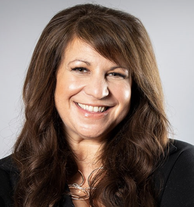 Smiling woman with long brown hair and a black blazer, posing for a professional portrait against a gray background.