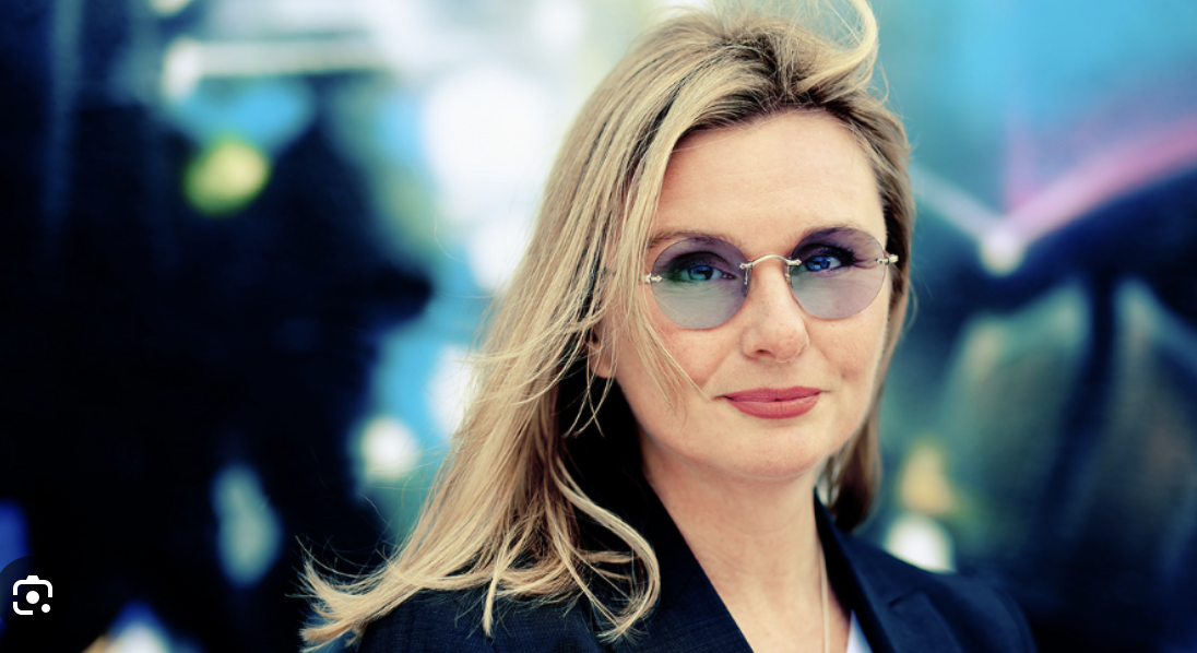 A woman with blonde hair wearing round sunglasses and a dark blazer, standing outdoors with a colorful blurred background.