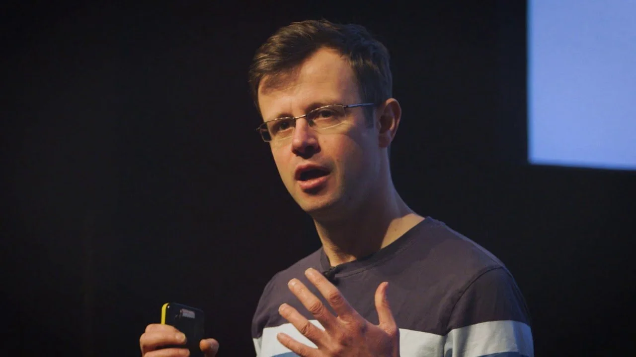 A man wearing glasses and a striped shirt is speaking, holding a remote or phone in his right hand, gesturing with his left hand.