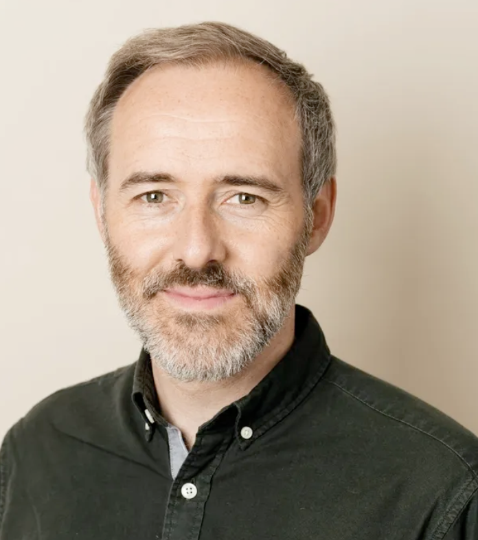Headshot of a middle-aged man with gray hair and beard, wearing a black button-up shirt, smiling slightly, against a light background.