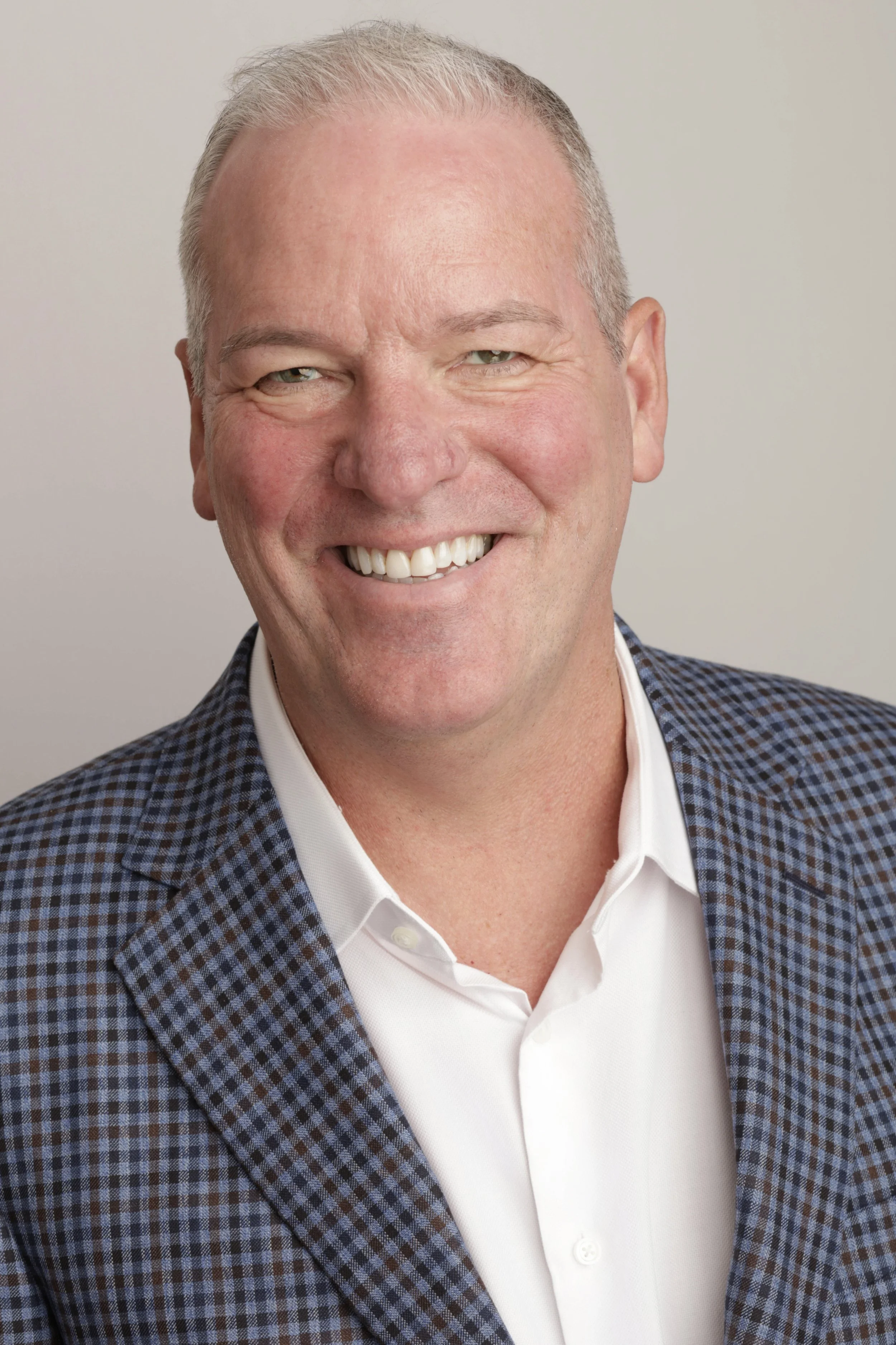 A smiling man with light-colored hair dressed in a white shirt and a checkered blazer.