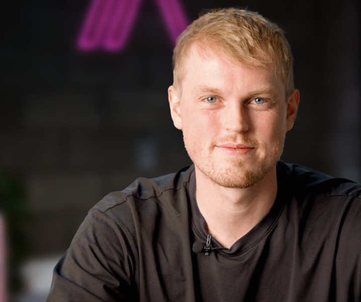 A young man with short blonde hair and blue eyes, wearing a black shirt, smiling at the camera.