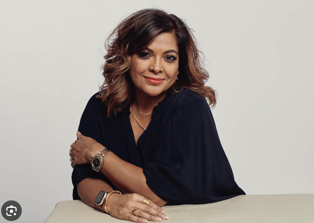 A woman with shoulder-length brown hair and light makeup, wearing a navy blue blouse, gold jewelry, and a watch, smiling and leaning on a table against a plain light gray background.