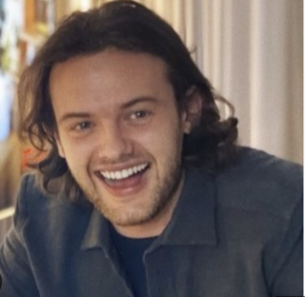 Young man with long, dark hair and light beard smiling and looking at the camera indoors.