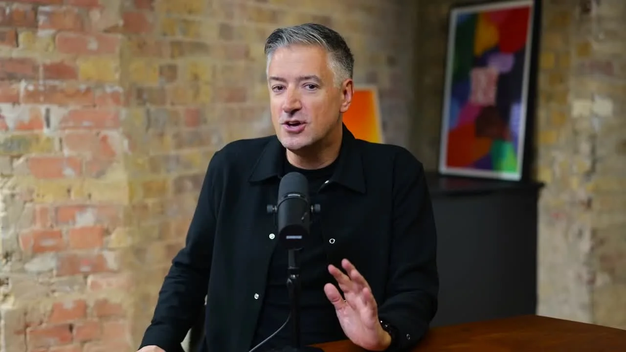 A man with gray hair speaking into a microphone during a talk or interview, sitting at a table with a brick wall and colorful artwork in the background.