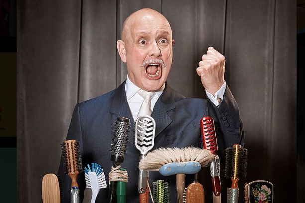 A man with a bald head in a suit is making a fist and shouting, standing behind a table with various hairbrushes and grooming tools.