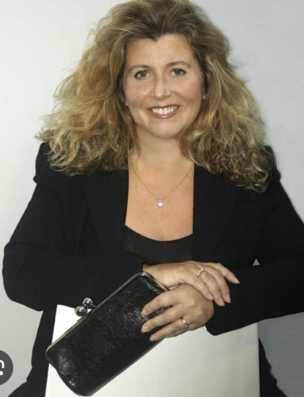 A woman with curly blonde hair smiling and holding a black clutch purse, dressed in a black blazer and top, against a plain background.