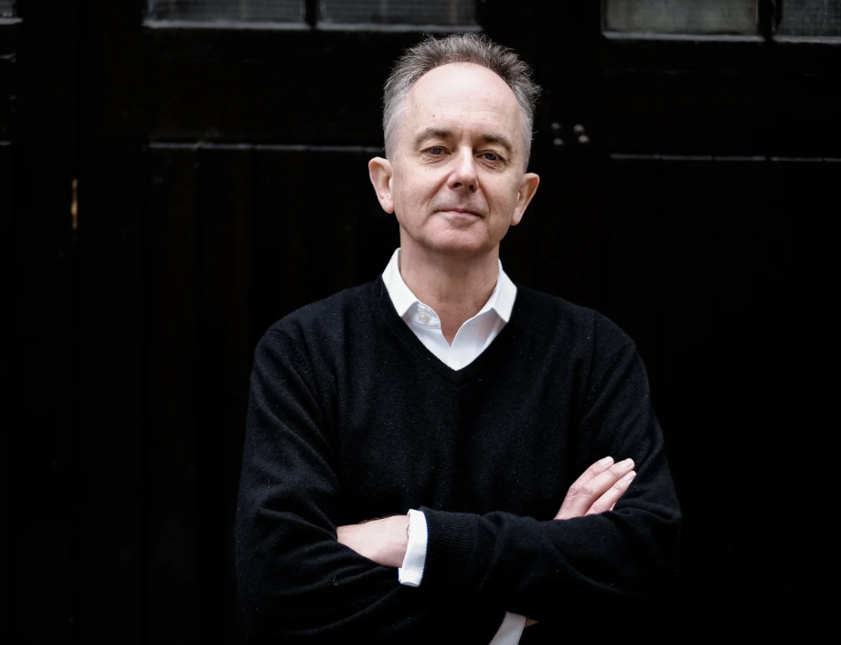 A middle-aged man with short gray hair wearing a white collared shirt and black sweater with arms crossed standing in front of a black wooden fence.