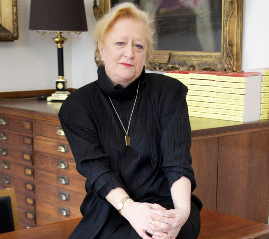 An older woman with short blonde hair, wearing a black blouse, sitting on a wooden chair in a room with a wooden dresser, a lamp, and a stack of books in the background.