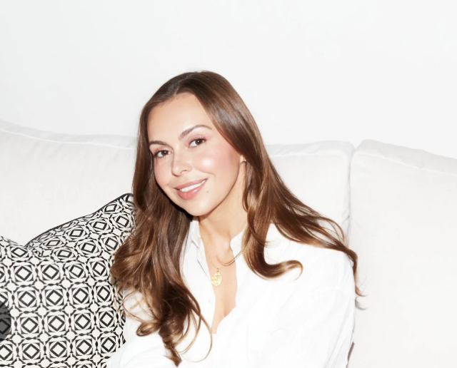 A woman with long brown hair, sitting on a white couch, smiling at the camera, wearing a white shirt, with patterned pillow behind her.
