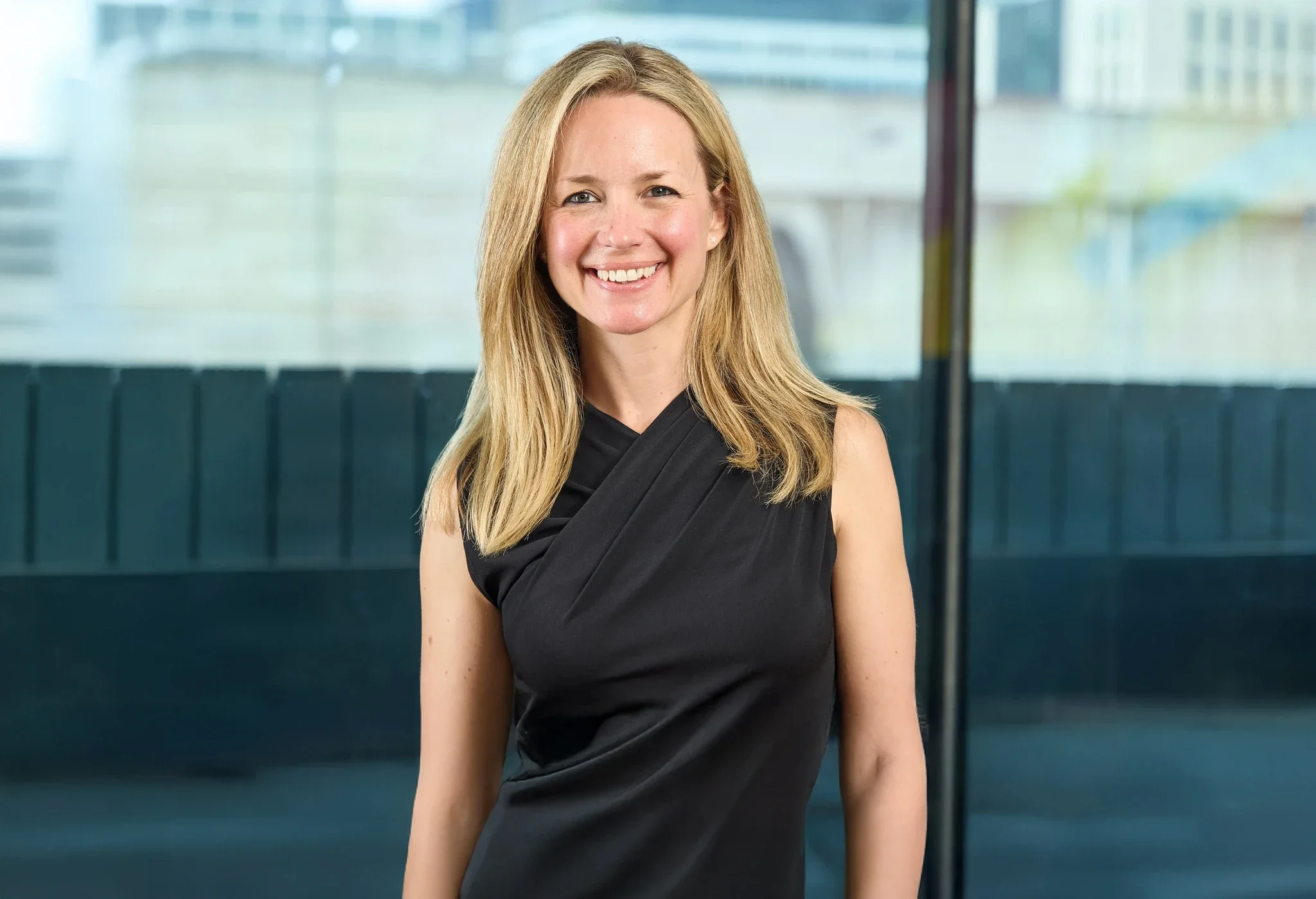 A smiling woman with blonde hair wearing a black sleeveless dress standing in front of a glass building.