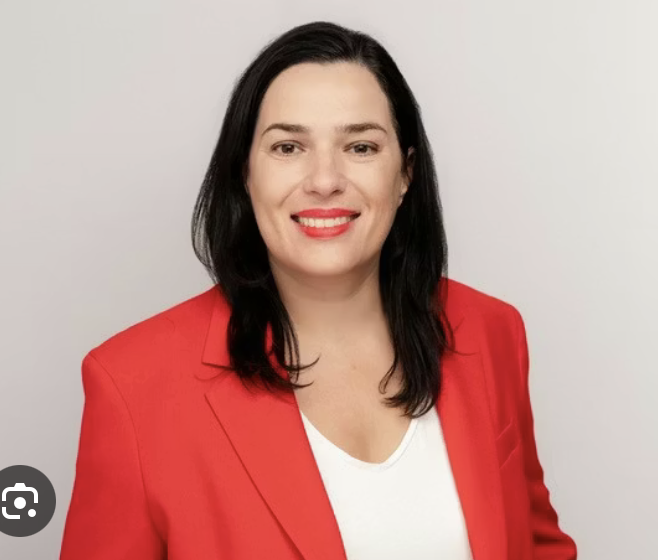 Professional woman with long dark hair, wearing a red blazer and white top, smiling against a plain light background.