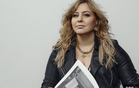 A woman with wavy blonde hair wearing a black leather jacket and gold jewelry, holding a newspaper, standing against a plain light gray background.