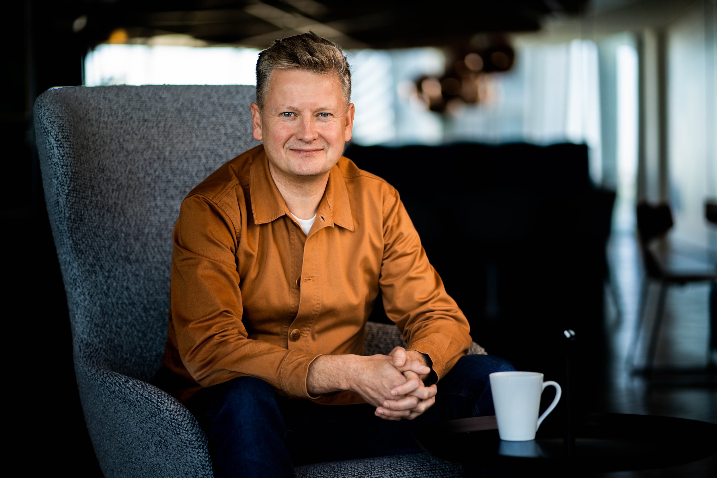 A middle-aged man with short, gray hair, wearing a brown shirt, sitting on a gray armchair with a white coffee mug on the table beside him in a cozy, well-lit indoor space.