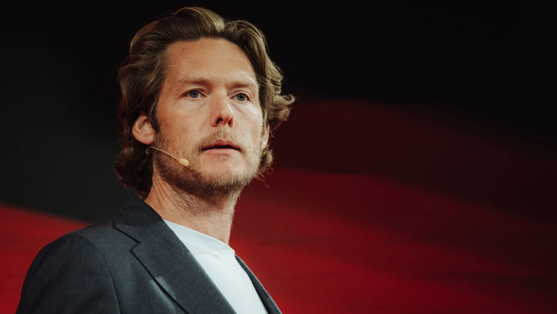 A man with wavy brown hair and a beard, wearing a dark suit jacket and a white shirt, speaking with a microphone headset on, against a red and black background.