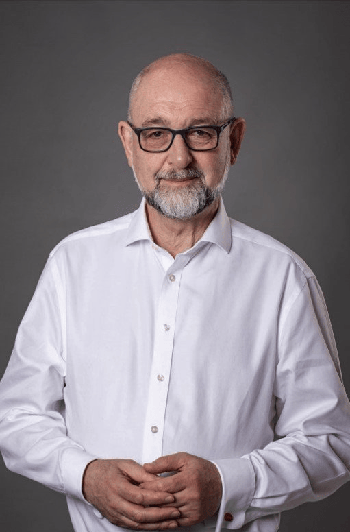 An older man with glasses, a beard, and a bald head wearing a white button-up shirt, standing against a gray background.