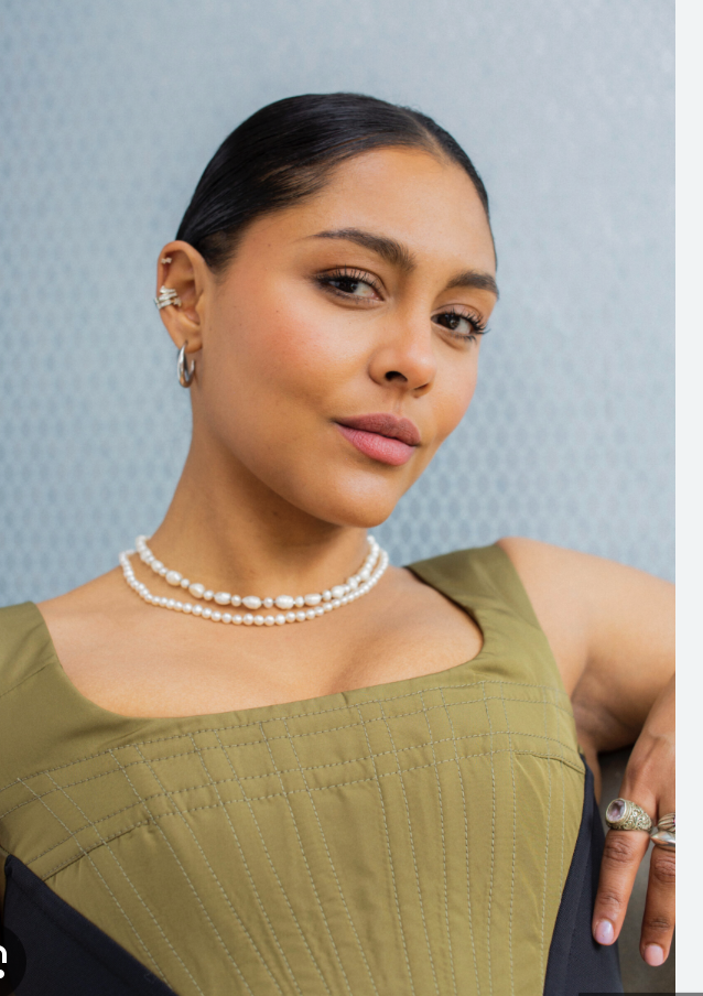 A woman with dark hair pulled back, wearing pearl jewelry and an olive green top, posing against a light blue background.