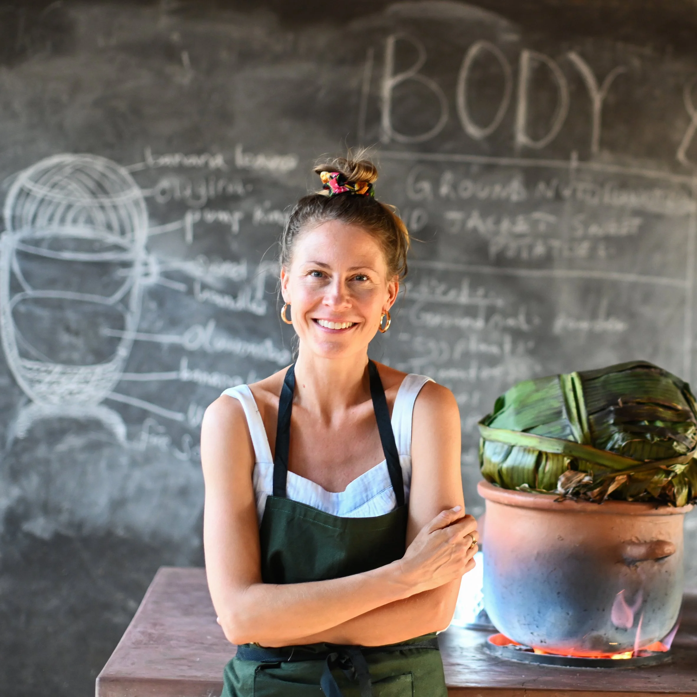 Lächelnde Frau in Kochschürze steht vor einem schwarzen Brett mit wissenschaftlichen Diagrammen und Texten, neben einer großen Tontopf mit an einem Herd erhitztem Bananenblatt.