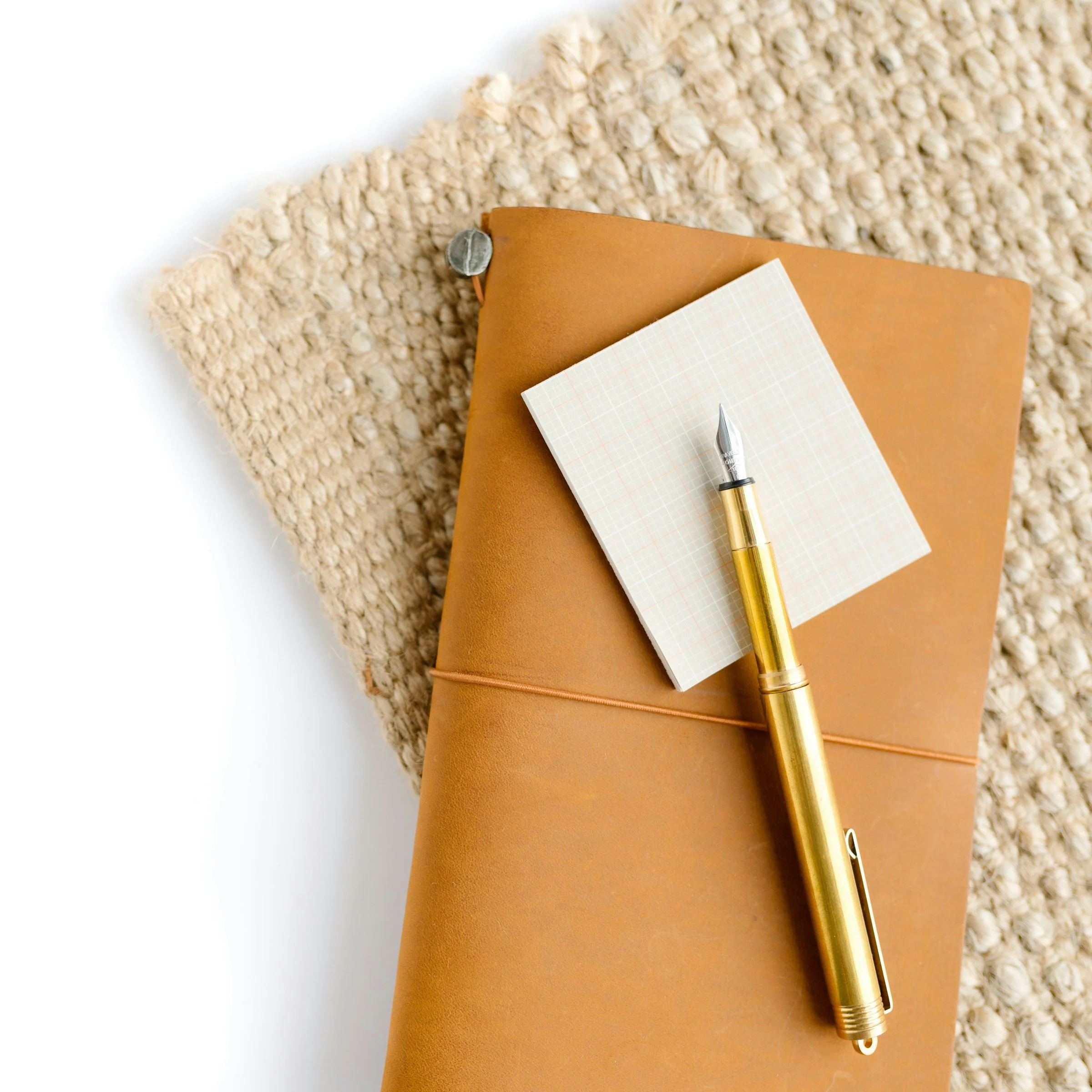 A leather-bound journal, a gold pen, a small paper notepad with grid lines, resting on a textured beige rug with a fluffy, woven appearance.