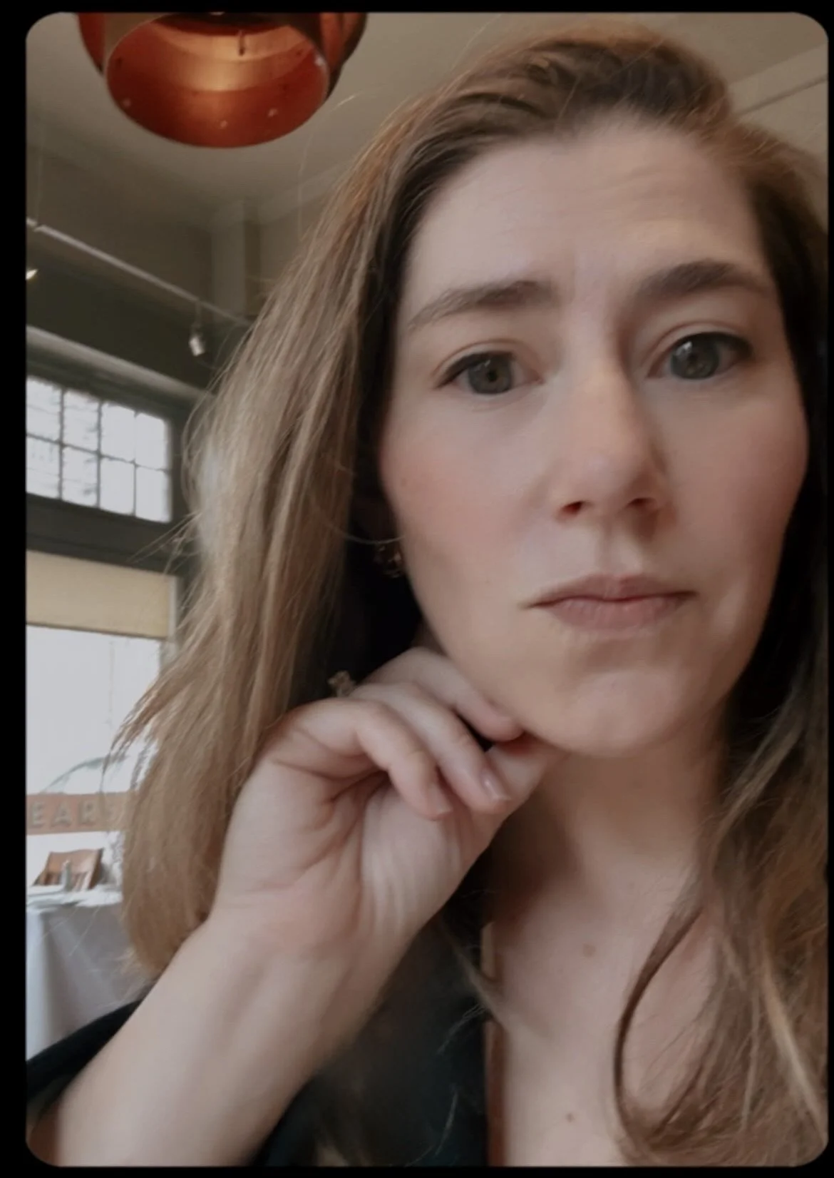 A close-up selfie of a woman with light skin, long light brown hair, and grey eyes, resting her chin on her hand inside a well-lit indoor space with large windows and industrial decor.