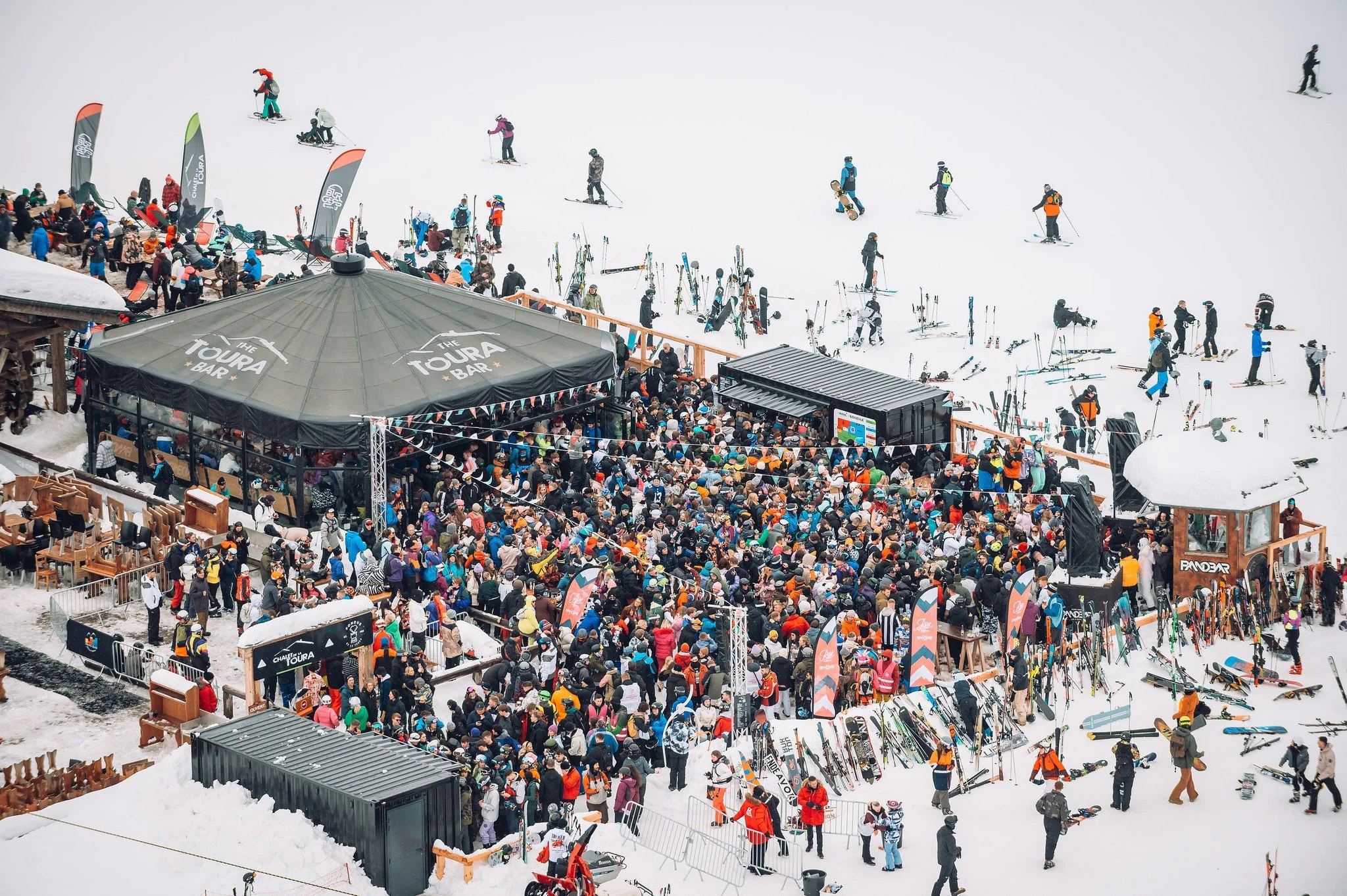 Foule de skieurs et de spectateurs au pied d'une station de ski enneigée avec un chapiteau et des stands de location de skis.