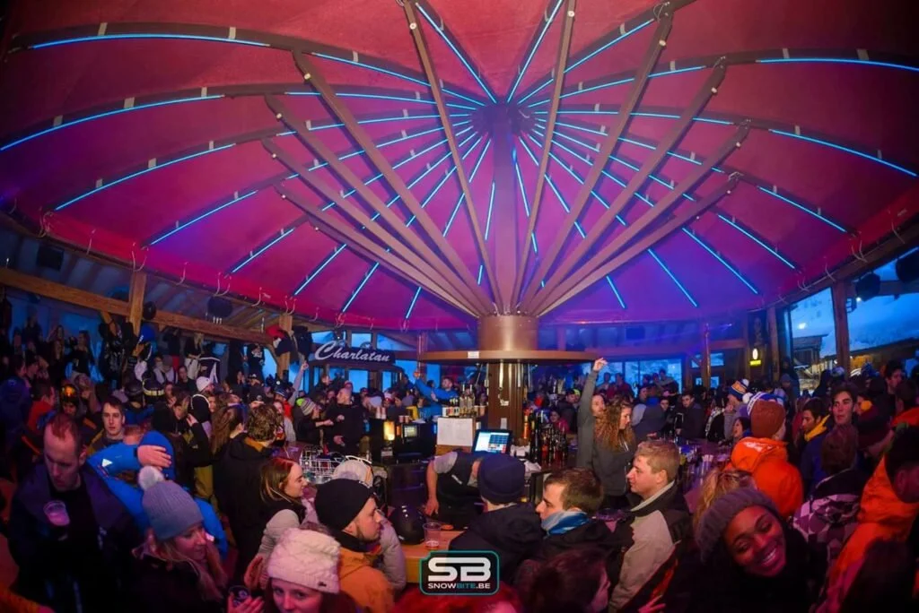 Une fête animée dans un espace intérieur avec un plafond en forme de parasol illuminé de néons bleus, beaucoup de personnes avec des vêtements d'hiver, un bar central, ambiance festive et lumineuse