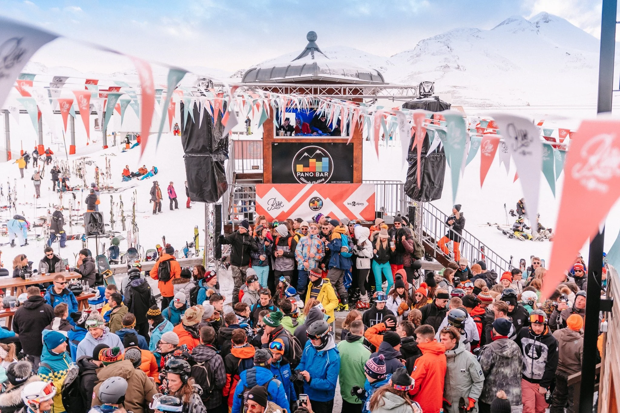 Foule de personnes en tenues de ski rassemblées devant un stand sur une station de ski enneigée, décorée avec des fanions colorés.