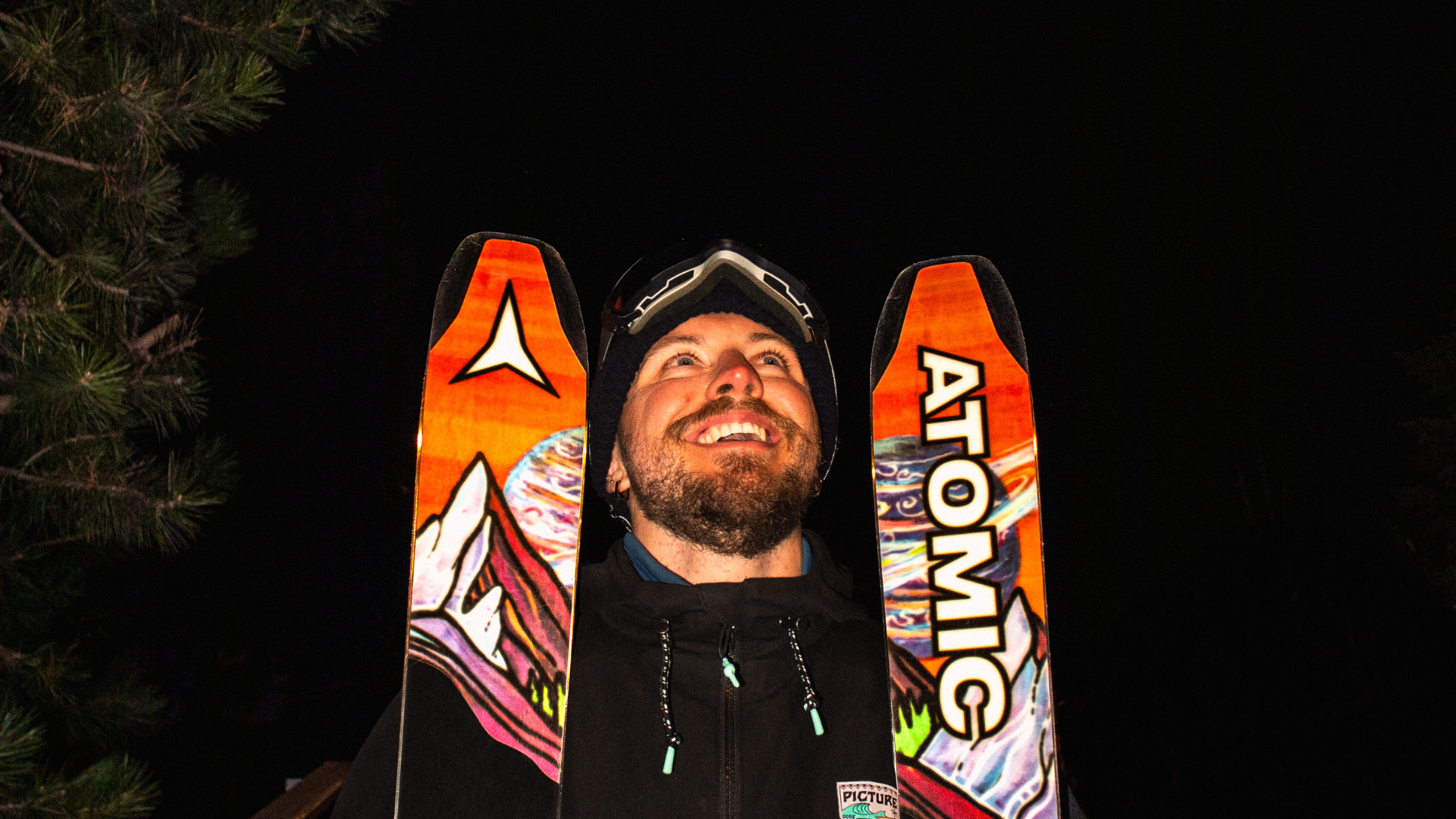 Jeune homme souriant portant un casque de ski, avec ses skis orange derrière lui, en bas d'une montagne, la nuit.