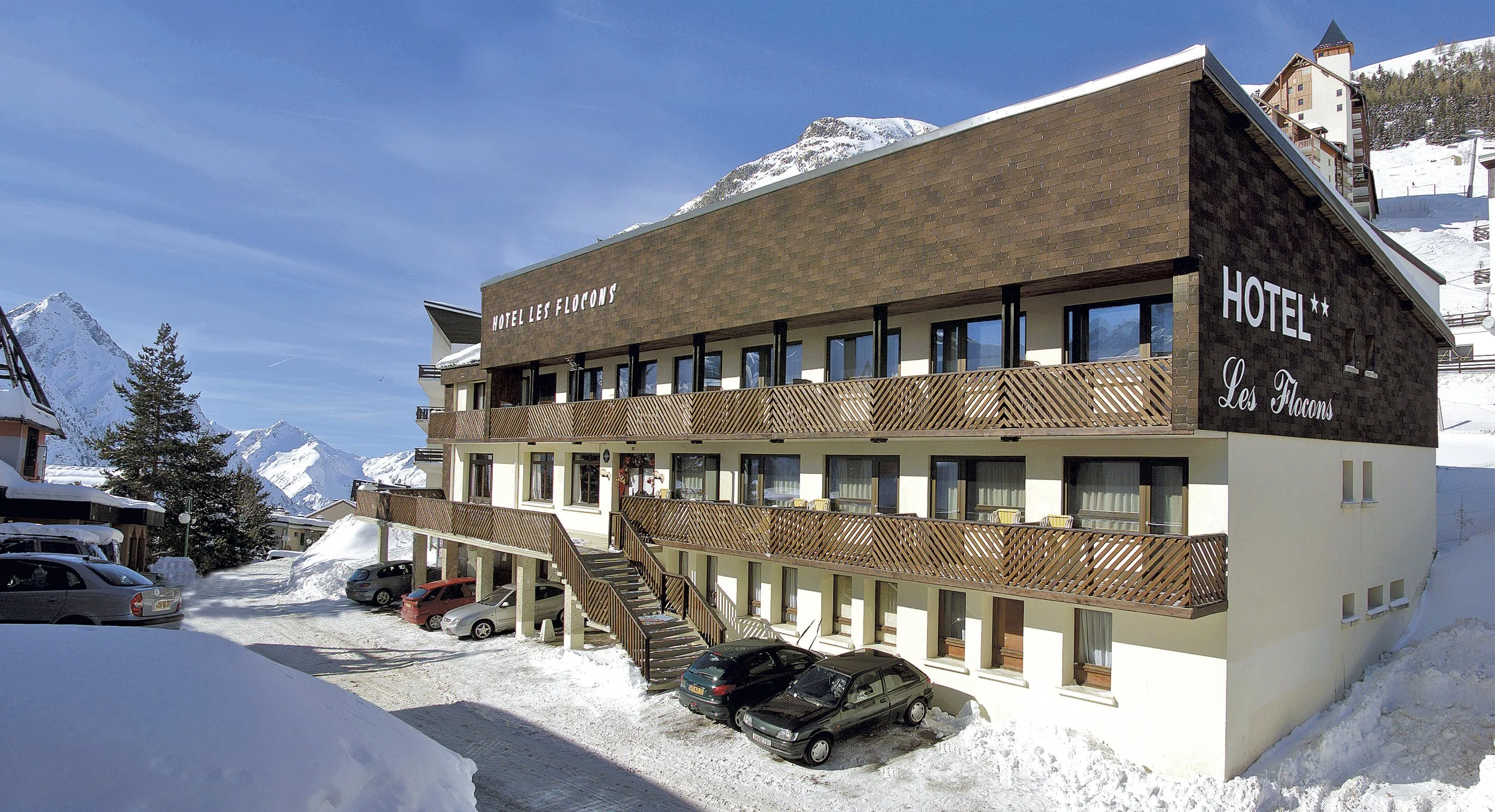 Un hôtel en montagne enneigée avec plusieurs voitures garées devant, et des montagnes en arrière-plan.