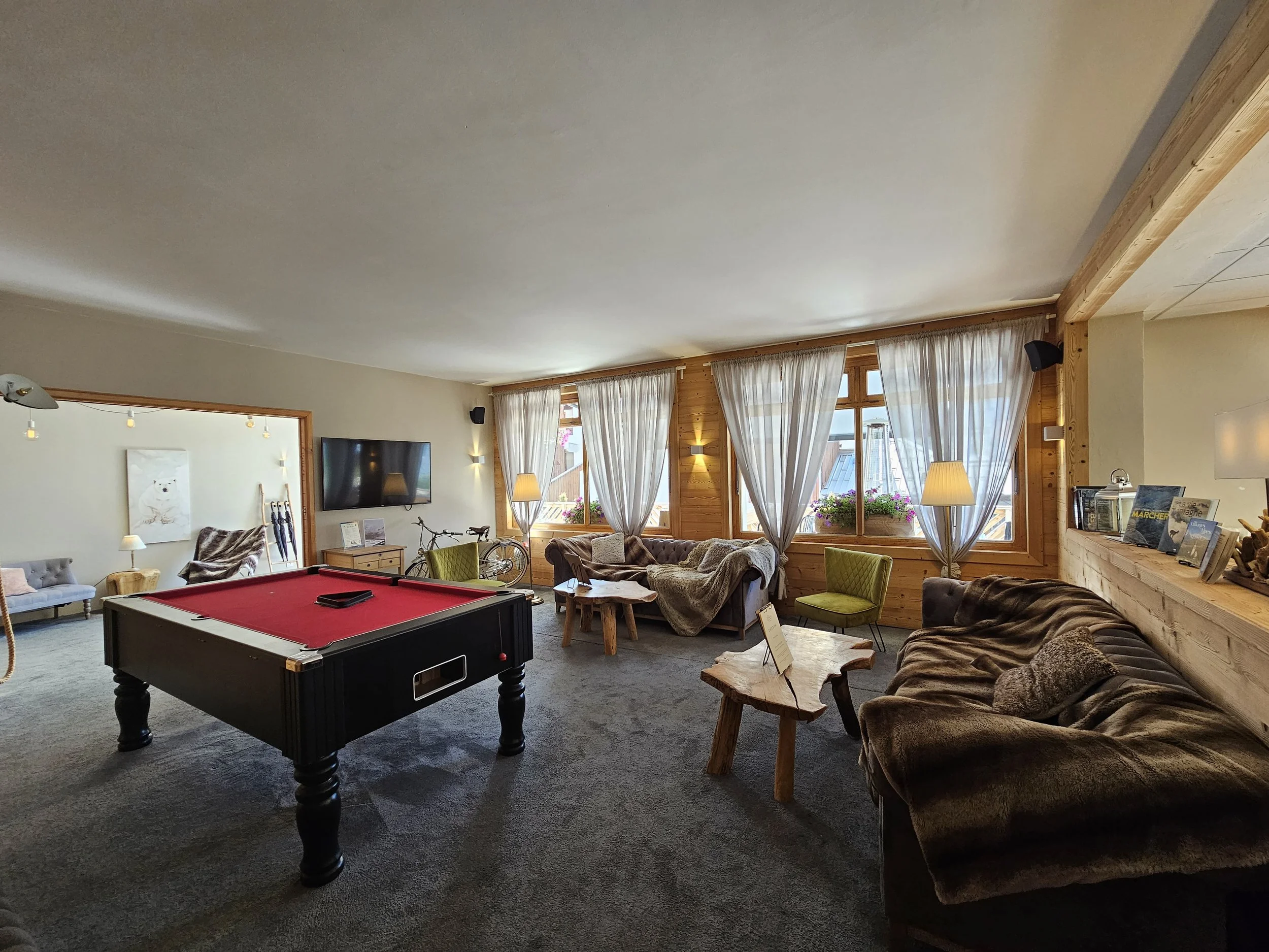 Salle de jeux avec un billard noir, plusieurs sofas en tissu, un mur avec une télévision, des fenêtres avec rideaux blancs, un vélo, des lampes, et des étagères avec des livres.