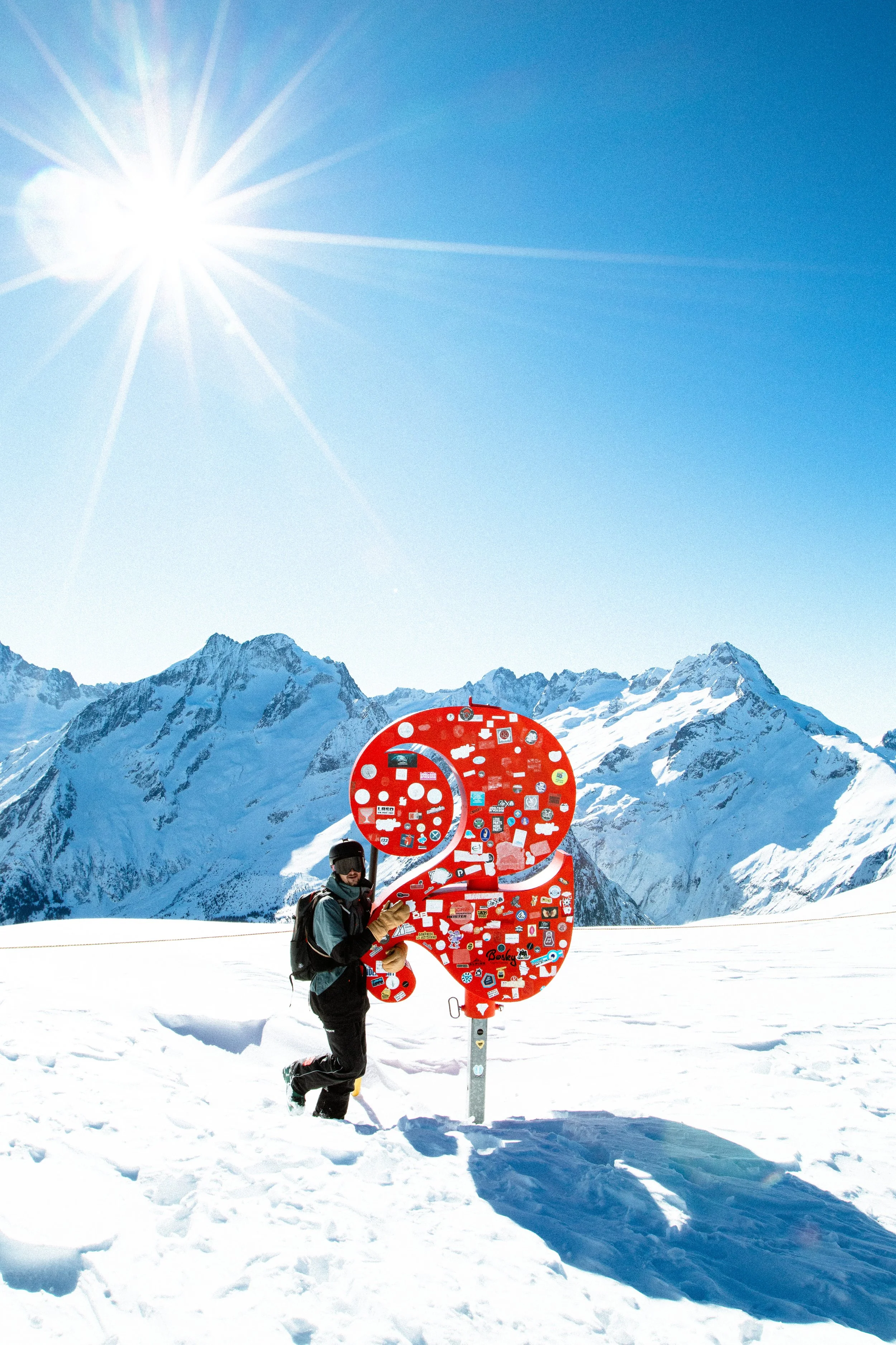 Un skieur en hiver, vêtu de vêtements chauds, se tient à côté d'un panneau rouge en forme de chiffre 23, dans un paysage montagneux enneigé avec un ciel clair et ensoleillé.