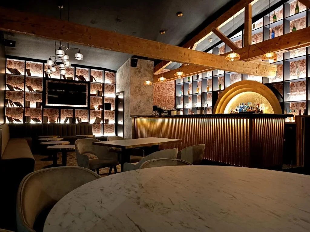 Intérieur d'un bar ou restaurant moderne avec bar en bois, étagères décoratives avec bouteilles, canapé beige, tables et chaises, éclairage chaleureux.