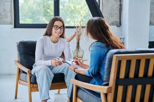 Two women sitting on chairs facing each other engaged in a conversation, one holding a smartphone and the other taking notes.