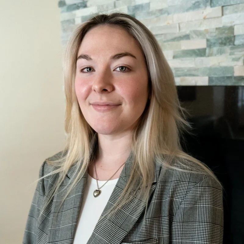 A woman with blonde hair wearing a plaid blazer and white top, standing indoors near a stone accent wall.