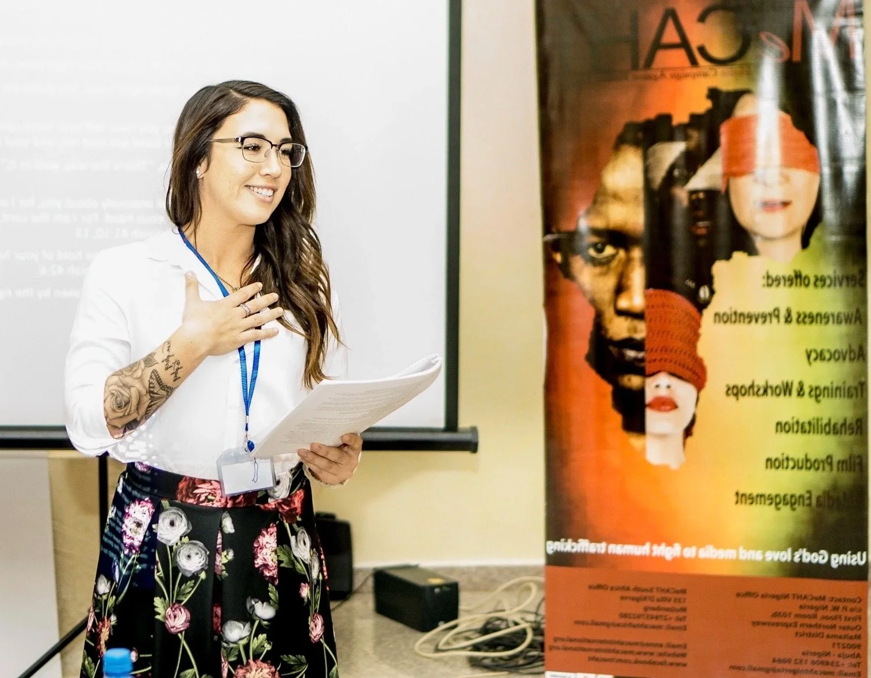 A woman with glasses, tattooed arm, and floral skirt smiling and speaking, holding papers, standing next to a colorful illustrated poster at a presentation or seminar.