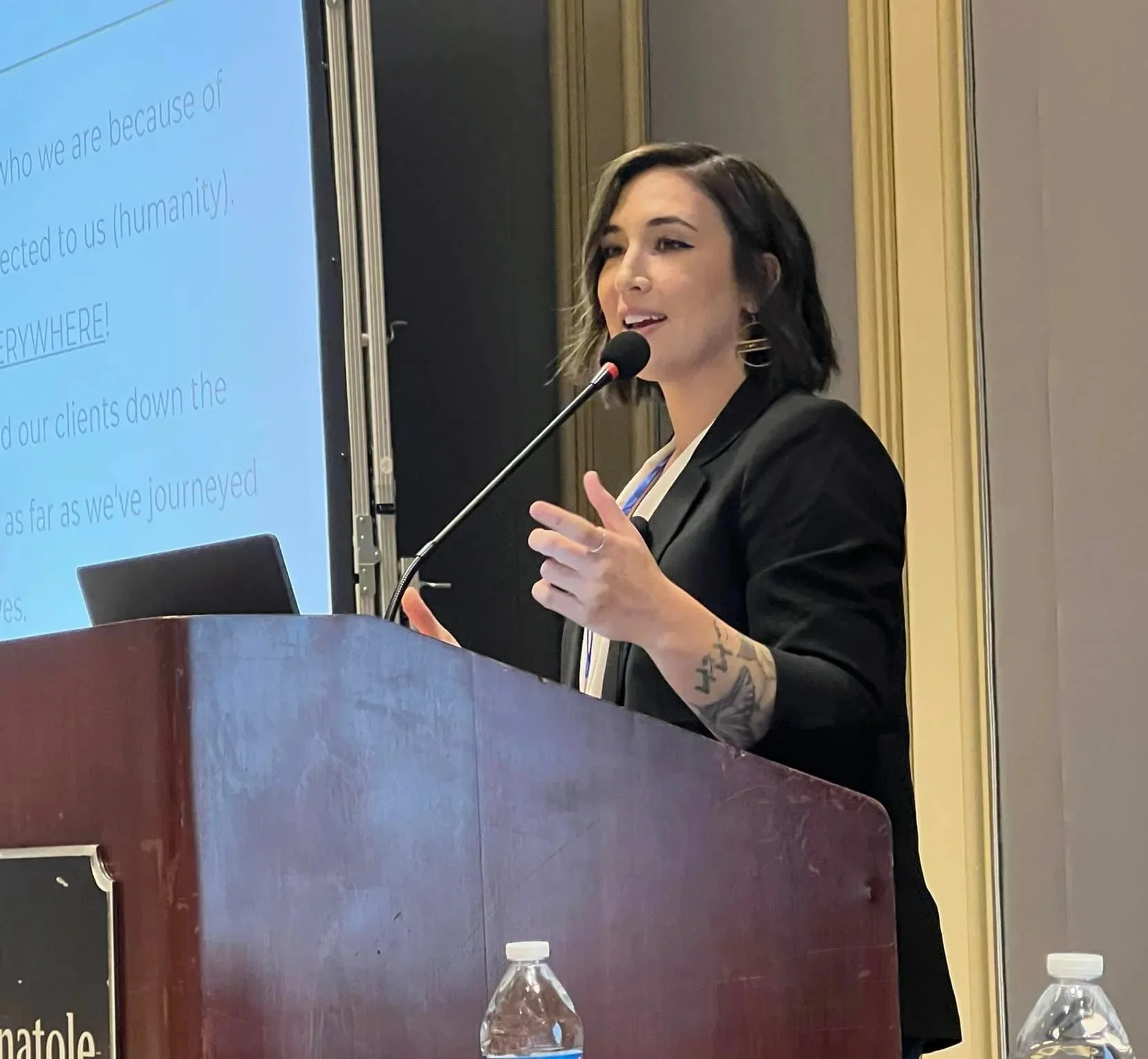 A woman with short black hair and earrings giving a presentation at a conference, standing behind a podium with a microphone. There is a large projection screen to her left with text displayed.
