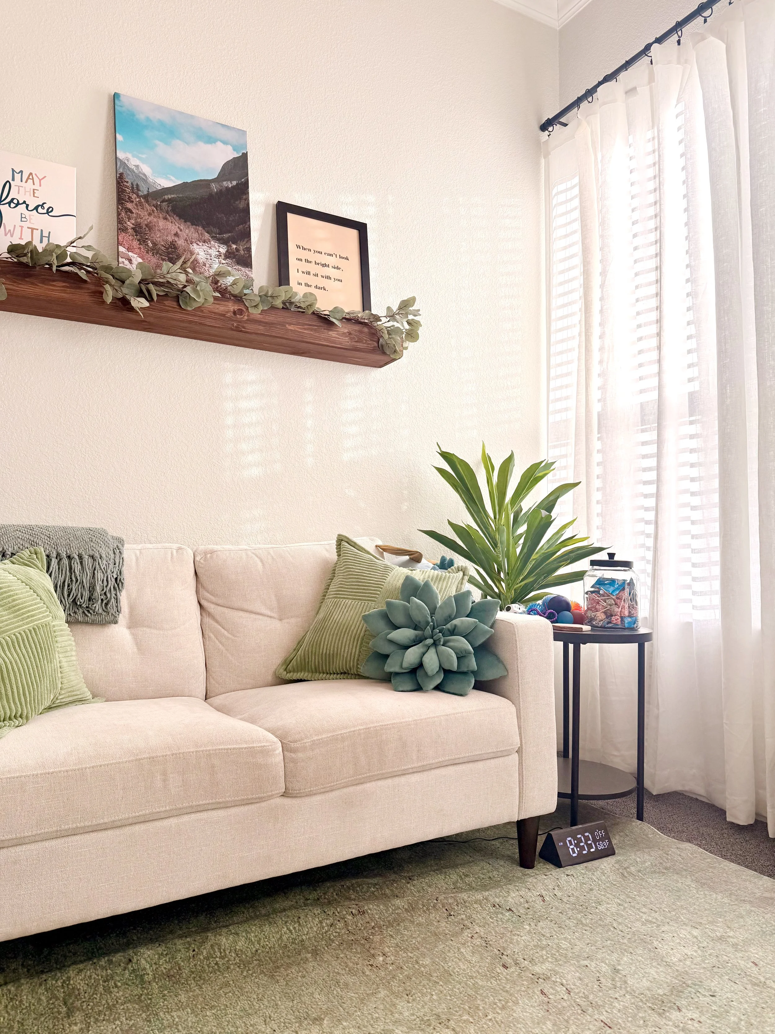 Living room with a beige sofa decorated with green pillows, a large green plant, a wooden shelf with framed pictures and inspirational quote, and a side table with a jar of colorful balls. Bright sunlight filters through white curtains.