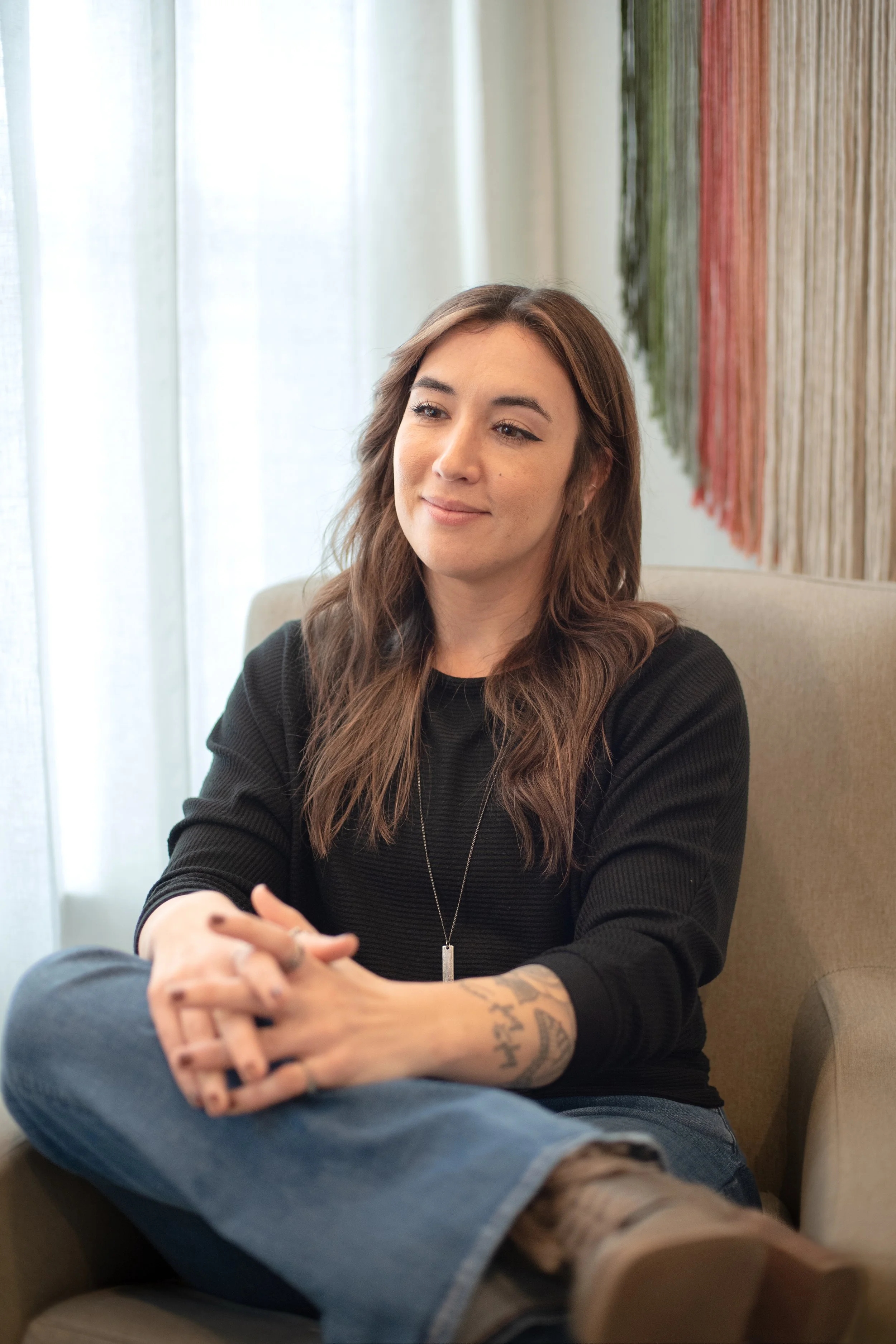 A young woman with brown hair and tattoos on her left arm sitting on a beige chair, wearing a black sweater, blue jeans, and beige boots, in a room with natural light and a textured wall behind her.
