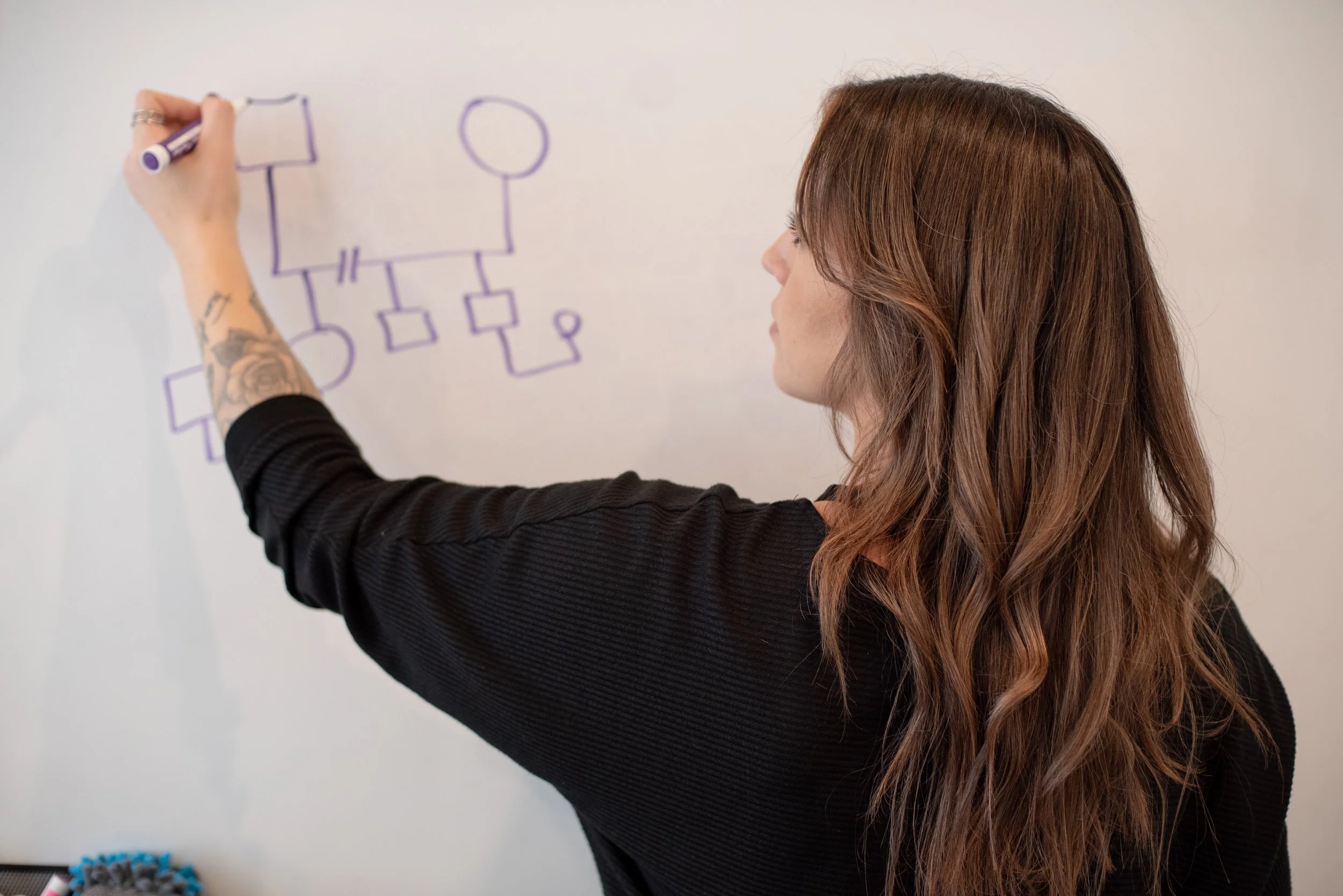 A woman with long, wavy brown hair drawing a circuit diagram on a whiteboard with a purple marker.