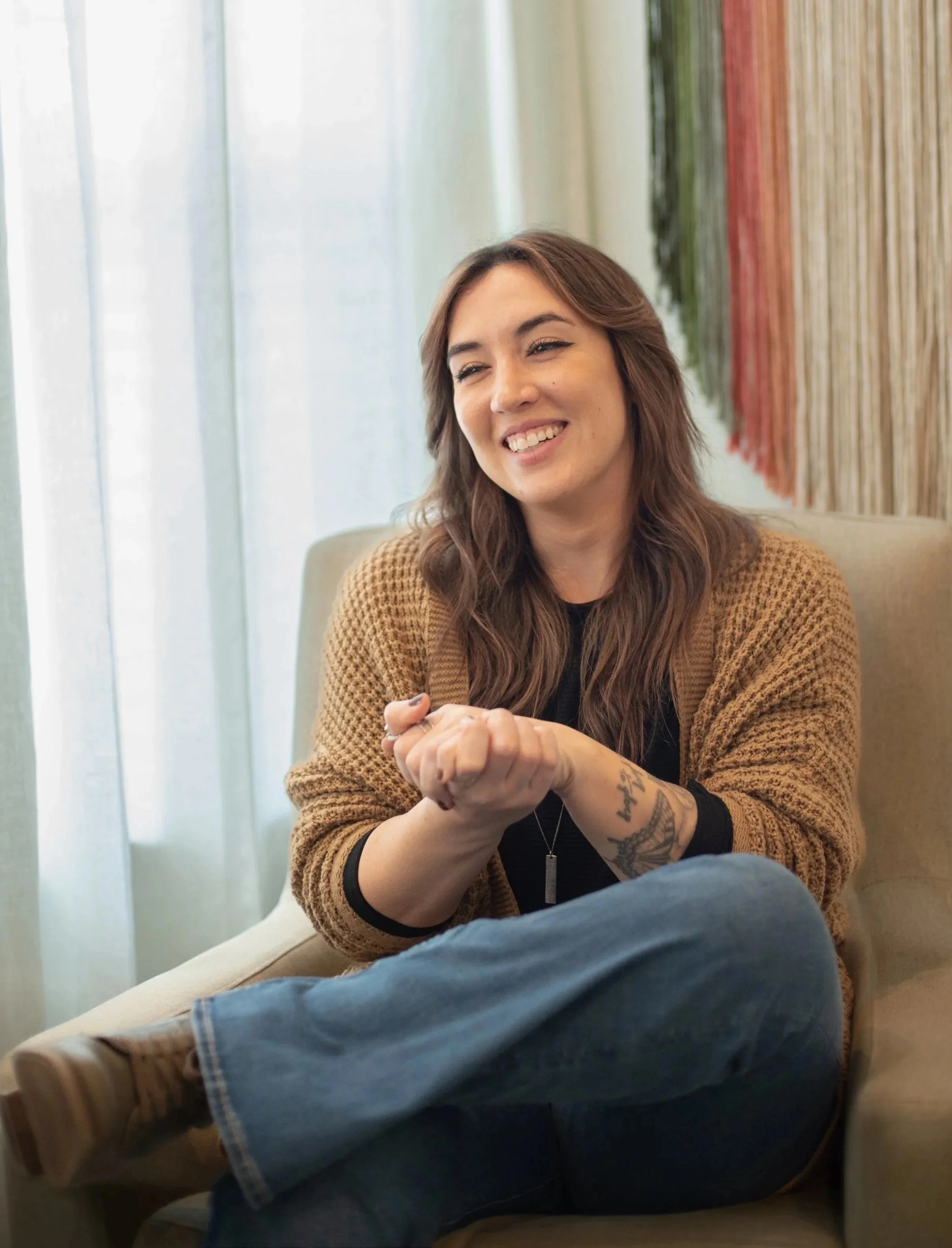 A woman with long brown hair sitting on a beige couch, smiling, wearing a brown cardigan over a black top, with crossed legs, in a room with sheer curtains and some wall hangings.