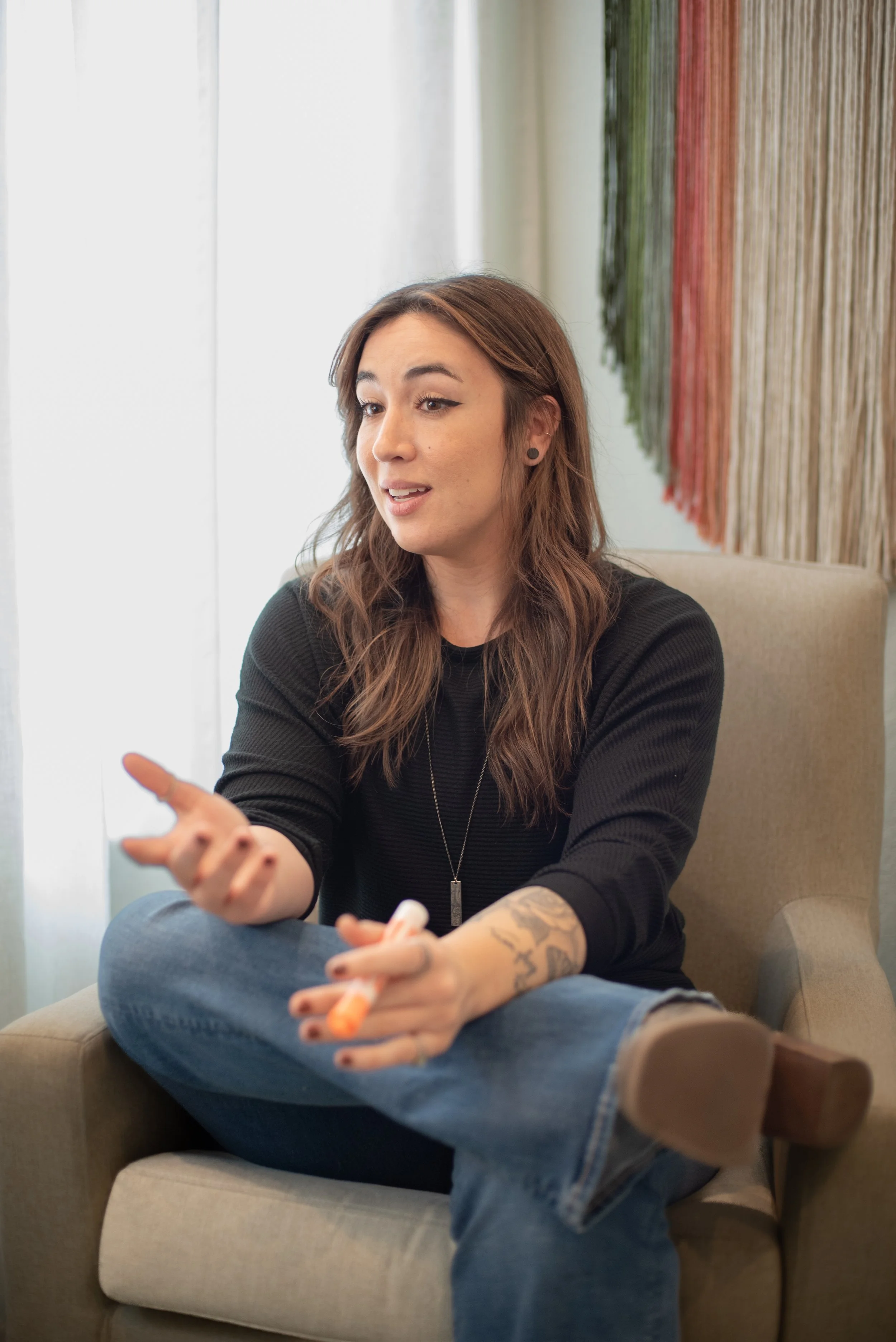 A woman with long brown hair, wearing a black sweater and jeans, sitting on a beige armchair, talking and gesturing with her hands, holding a tube of lipstick. Behind her is a colorful woven wall hanging.